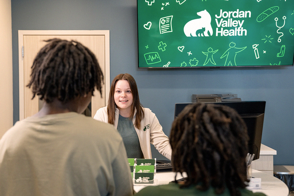 A Jordan Valley staff member greets two visitors at a clinic front desk.